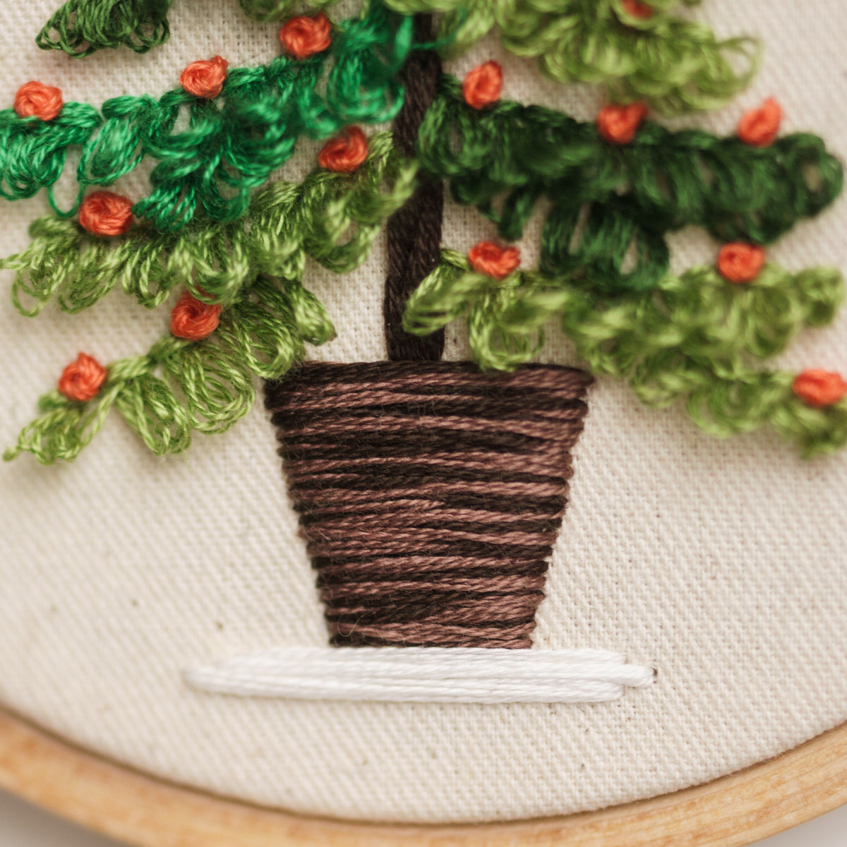 May include: Embroidered artwork of a potted plant with green leaves and orange flowers. The plant is in a brown pot with a white base. The artwork is framed in a wooden hoop.