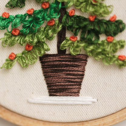 May include: Embroidered artwork of a potted plant with green leaves and orange flowers. The plant is in a brown pot with a white base. The artwork is framed in a wooden hoop.