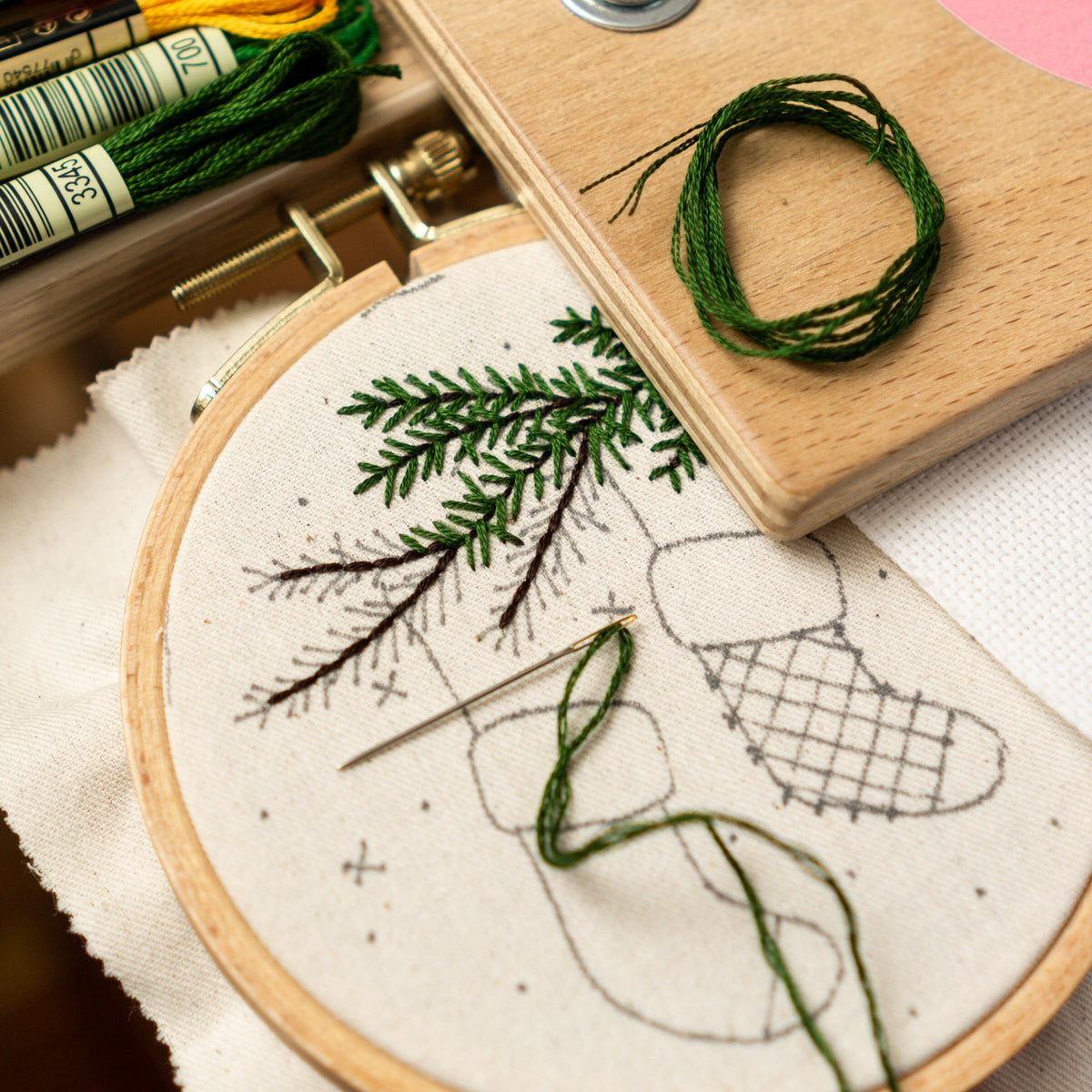 May include: A close-up of a partially completed embroidery project featuring a green pine branch and a boot. The embroidery hoop is made of wood and the fabric is white with a gray outline of the design. A needle and green embroidery floss are visib
