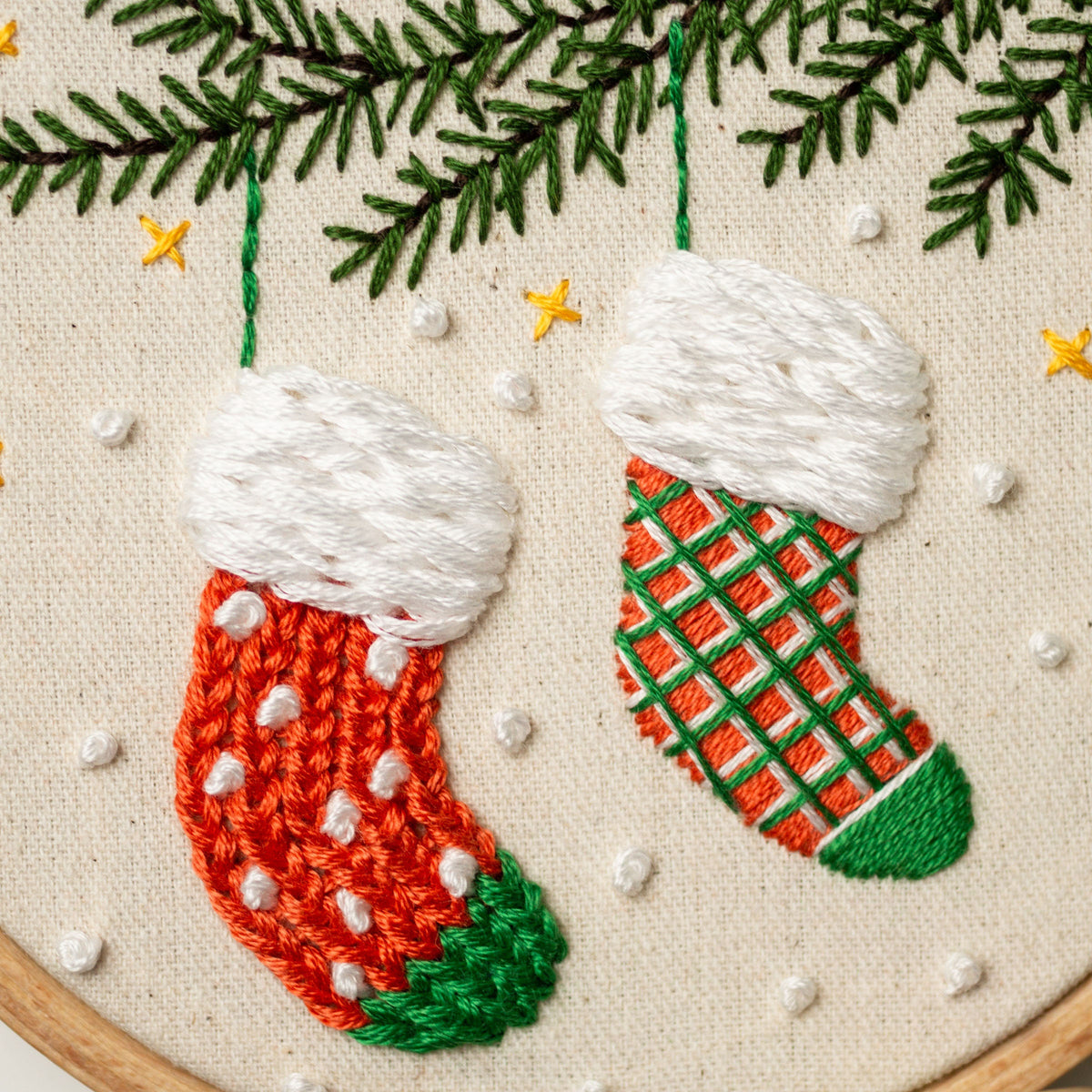 May include: Two embroidered Christmas stockings hanging from a green pine branch. One stocking is orange and green with white polka dots, the other is orange and green with a checkered pattern.