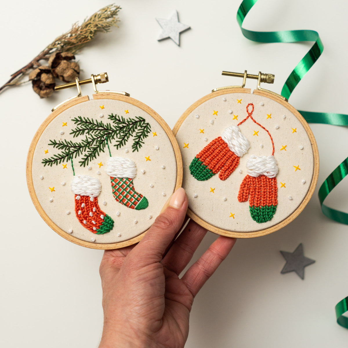 May include: Two embroidery hoops with Christmas-themed designs. One hoop features two red and green stockings hanging from a green pine branch. The other hoop features two red and green mittens with white cuffs.