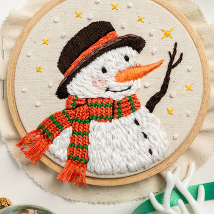 May include: Embroidered snowman with a brown hat, orange and green scarf, and white yarn body. The snowman is surrounded by white dots on a beige background.
