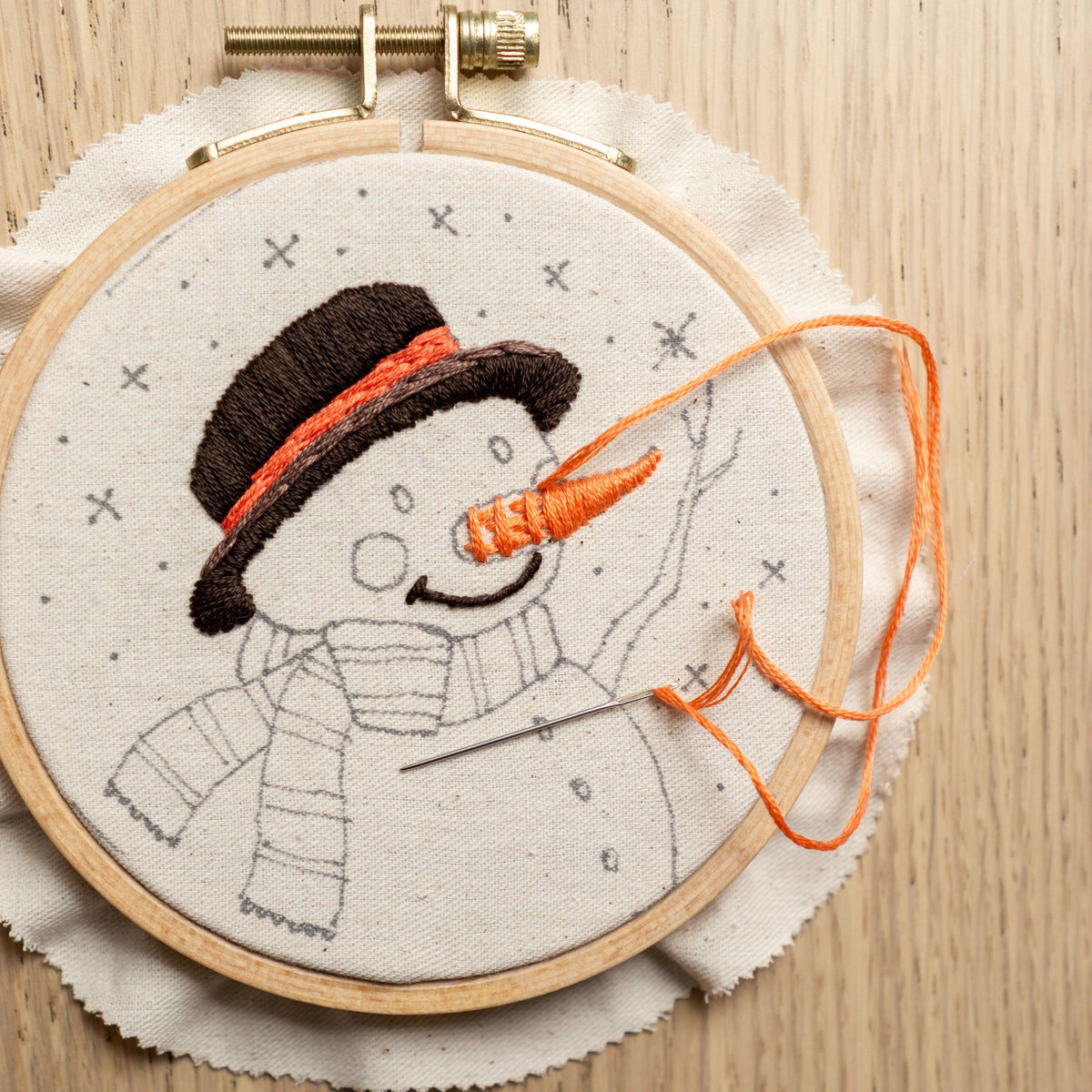 May include: A close-up of a partially completed embroidery hoop with a snowman design. The snowman is wearing a black hat with an orange scarf and has a carrot nose. The embroidery floss is orange.