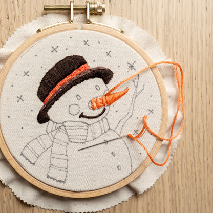 May include: A close-up of a partially completed embroidery hoop with a snowman design. The snowman is wearing a black hat with an orange scarf and has a carrot nose. The embroidery floss is orange.