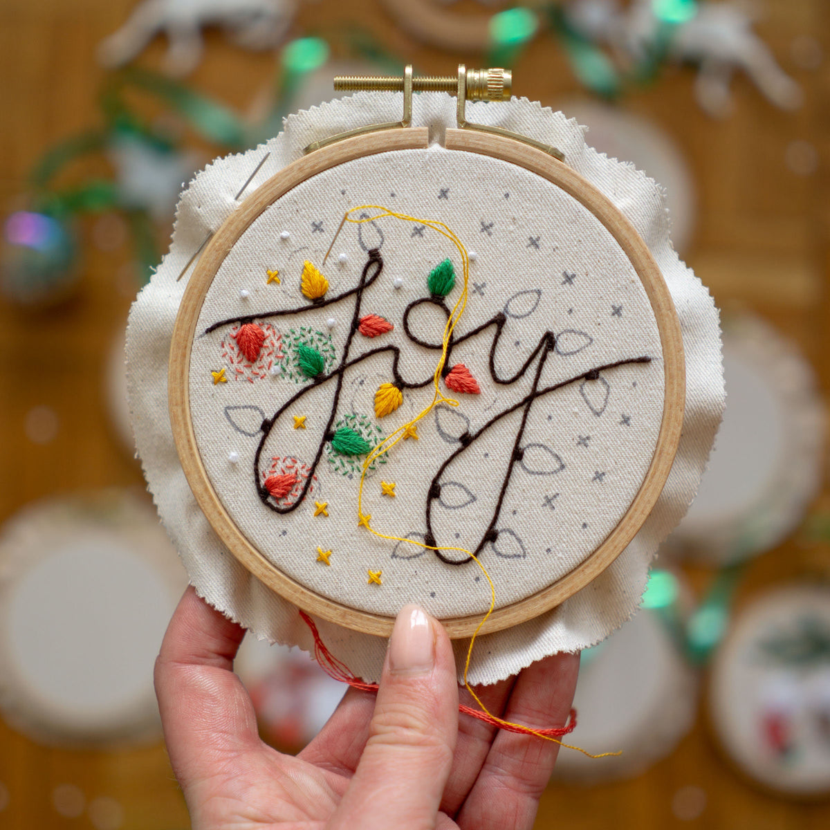 May include: Embroidered hoop art with the word "Joy" in black cursive lettering surrounded by colorful floral embroidery and yellow string lights.