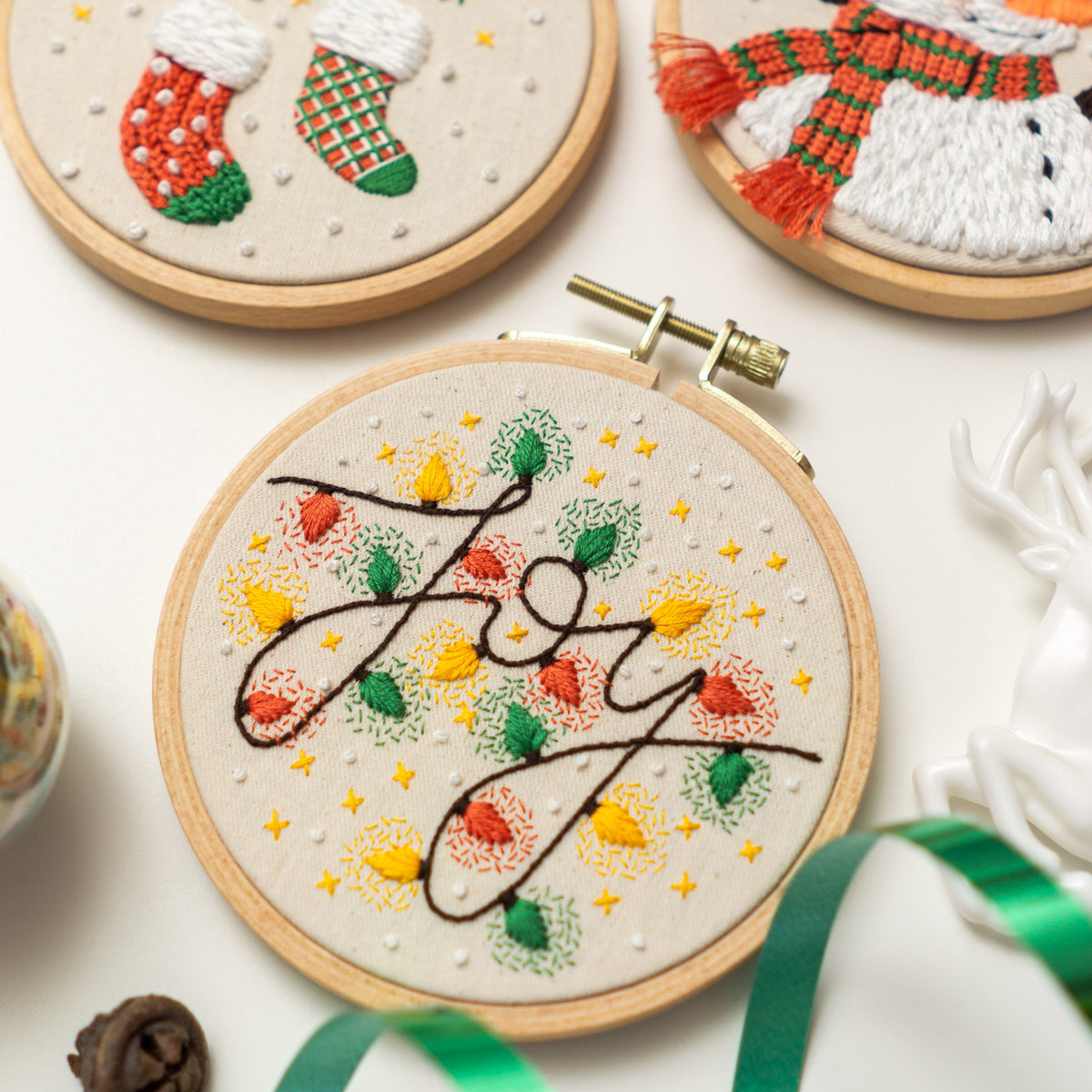May include: Embroidered hoop art with the word "Joy" in a cursive font surrounded by green leaves and yellow and orange lights. The background is a white fabric with small white dots.