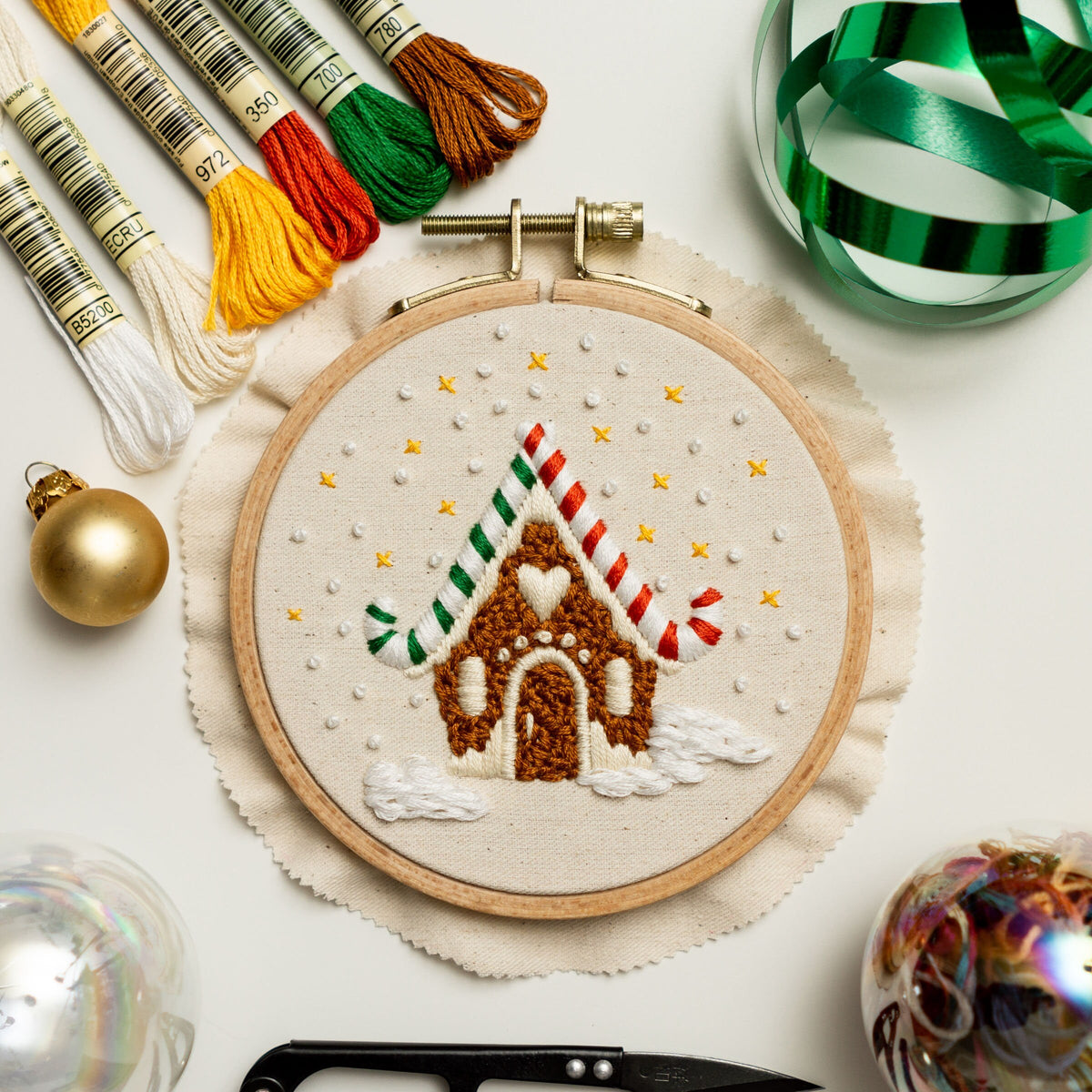 May include: A gingerbread house embroidery hoop with a white background and brown, red, and green embroidery floss. The hoop is surrounded by a white fabric with a ruffled edge.