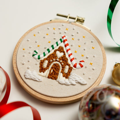 May include: A hand-stitched embroidery hoop featuring a gingerbread house with candy cane accents. The house is decorated with white and brown frosting and a heart-shaped window. The background is a white fabric with a scattering of white beads and 