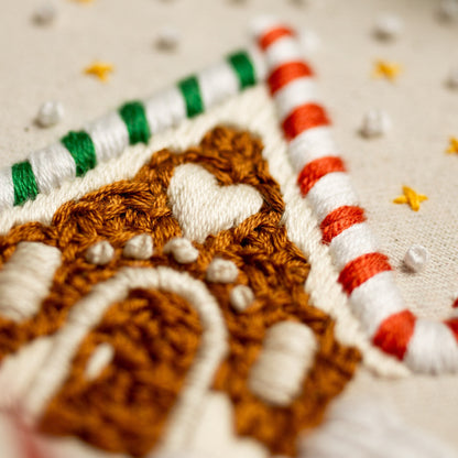 May include: Close-up of a gingerbread house embroidered on a white fabric. The house is brown with white icing and a heart-shaped window. The house is surrounded by a red and white candy cane.
