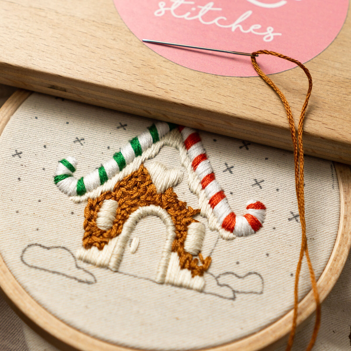 May include: A close-up of a gingerbread house embroidery pattern on a white fabric with brown, green, and red embroidery floss. The embroidery is in a hoop with a wooden frame.