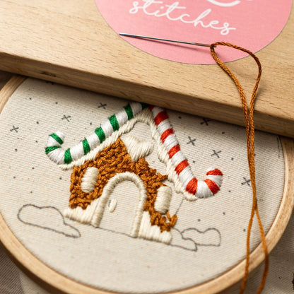 May include: A close-up of a gingerbread house embroidery pattern on a white fabric with brown, green, and red embroidery floss. The embroidery is in a hoop with a wooden frame.