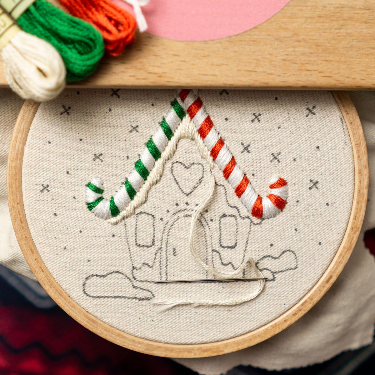 May include: A close-up of a white embroidery hoop with a gingerbread house design. The house is outlined in black and has red and green candy cane accents. The embroidery is partially complete, with white thread used to stitch the candy canes.