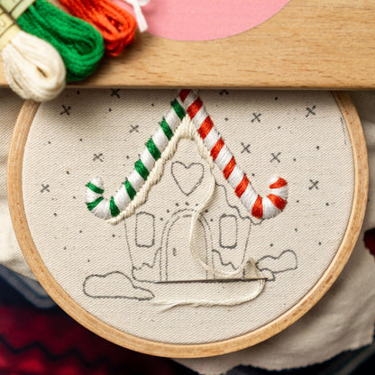May include: A close-up of a white embroidery hoop with a gingerbread house design. The house is outlined in black and has red and green candy cane accents. The embroidery is partially complete, with white thread used to stitch the candy canes.