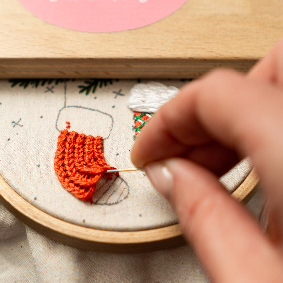 May include: Close-up of a hand embroidering a red knitted stocking on a white fabric with a wooden embroidery hoop. The stocking is partially embroidered with a green and white pattern.