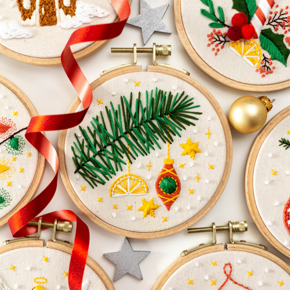 May include: A set of five embroidery hoops with various Christmas-themed designs. The designs include a pine branch with ornaments, a holly sprig with berries, a gingerbread house, and a candy cane. The hoops are all made of wood and have a natural 