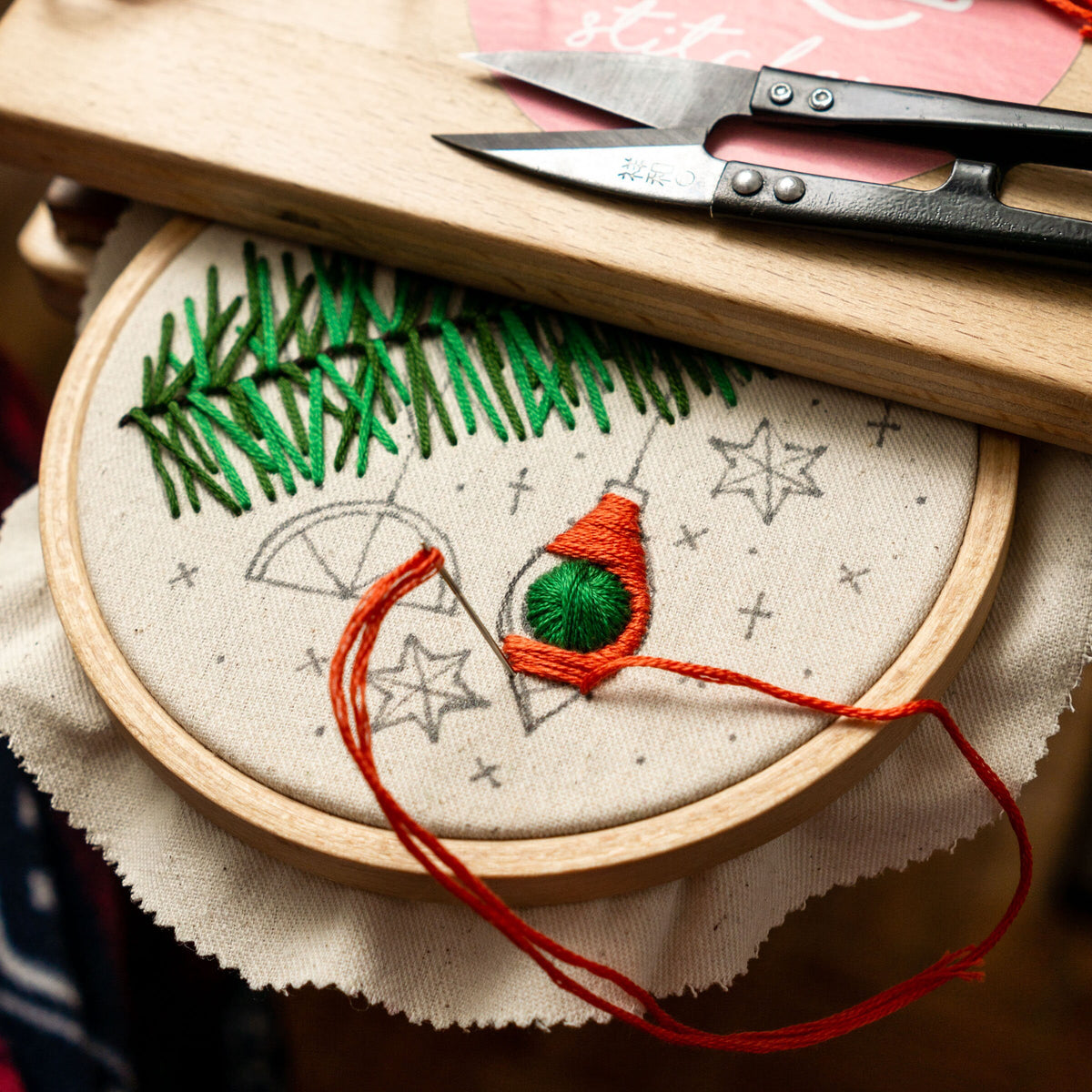 May include: A close-up of a partially completed embroidery project depicting a Christmas tree with ornaments. The embroidery hoop is made of wood and the fabric is white. The embroidery floss is red and green.