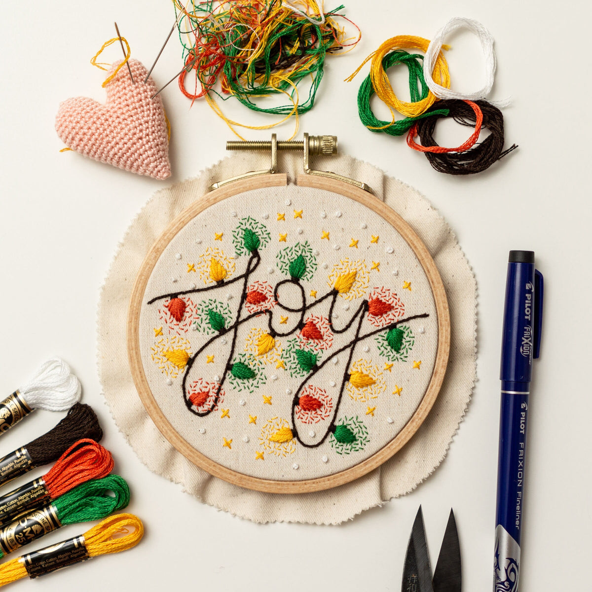 May include: A close-up of a white embroidery hoop with the word "Joy" stitched in colorful thread. The hoop is surrounded by embroidery floss, scissors, and a pen.