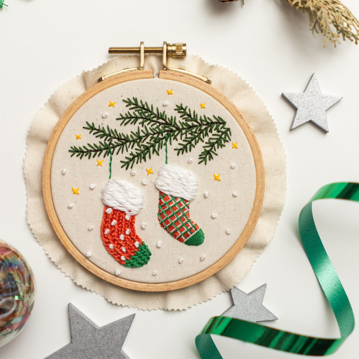 May include: Embroidered Christmas stocking ornament with a green pine branch and two red and green stockings hanging from it. The ornament is in a wooden embroidery hoop with a white fabric background.