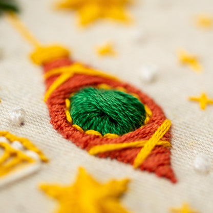 May include: Close-up of a red, yellow, and green embroidered ornament. The ornament is shaped like a teardrop and has a green center with yellow stitching around it. The ornament is surrounded by white fabric.