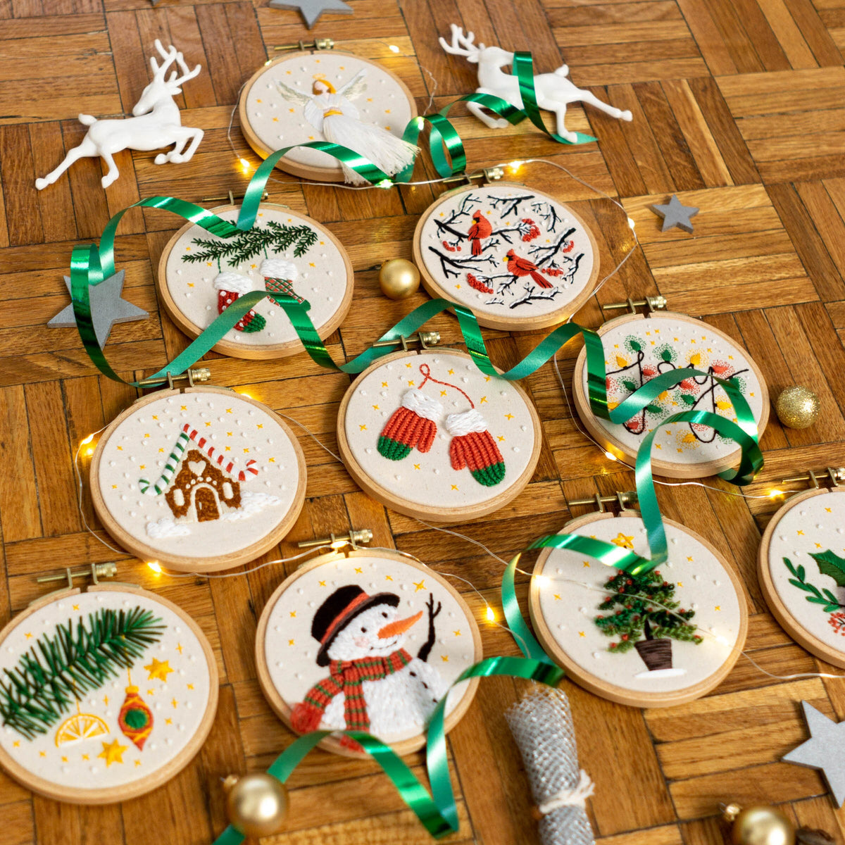May include: A collection of 10 round embroidery hoops with various Christmas-themed designs, including a snowman, a gingerbread house, a Christmas tree, and a pair of mittens. The hoops are arranged on a wooden surface with green ribbon and white li