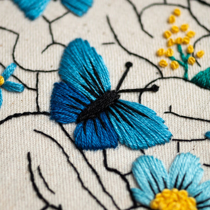 May include: Close-up of a blue butterfly embroidered on a white fabric with black thread. The butterfly has a black body and blue wings. The fabric has a black embroidered vine pattern.