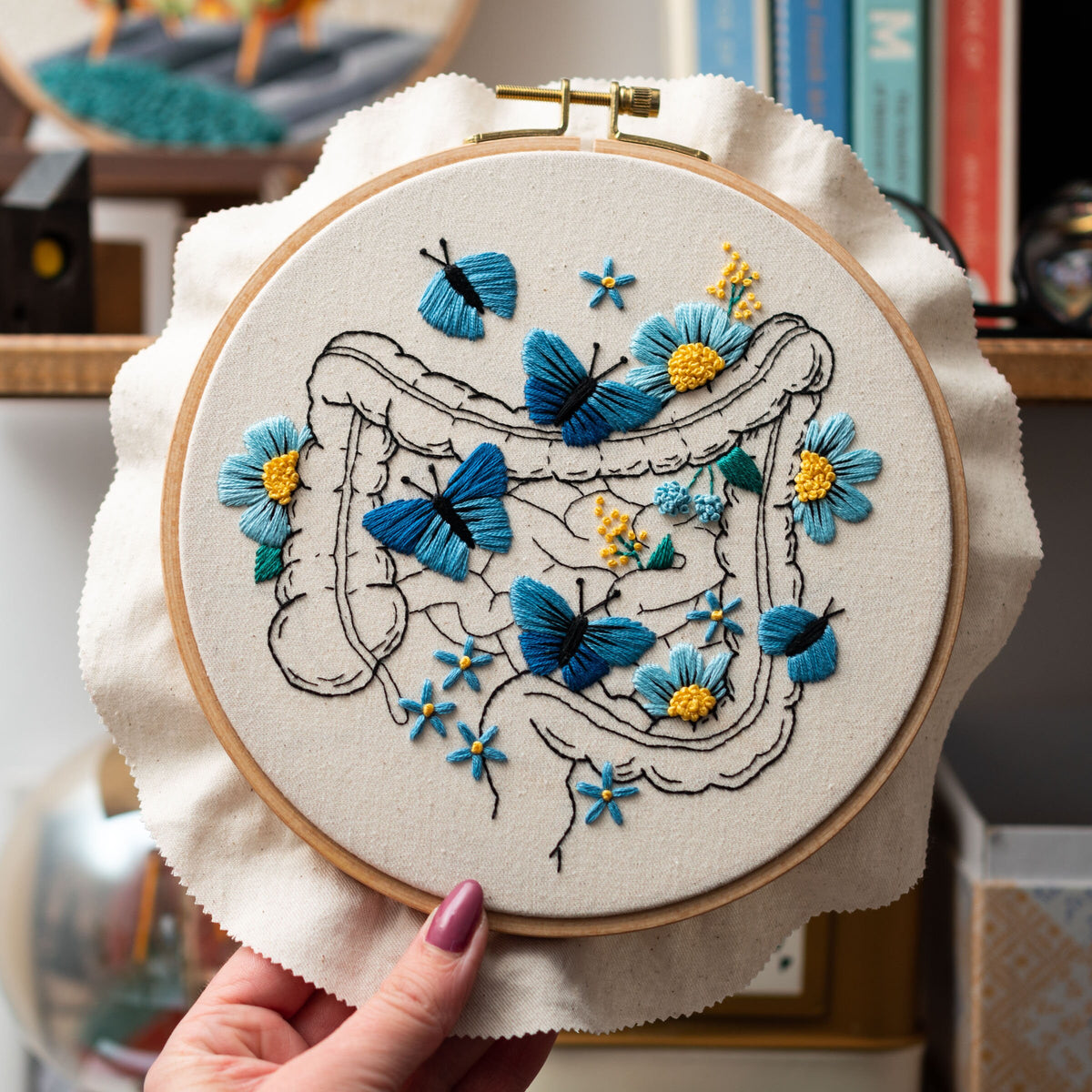 May include: Embroidered hoop art featuring a blue and yellow anatomical illustration of the human intestines with blue butterflies and flowers.