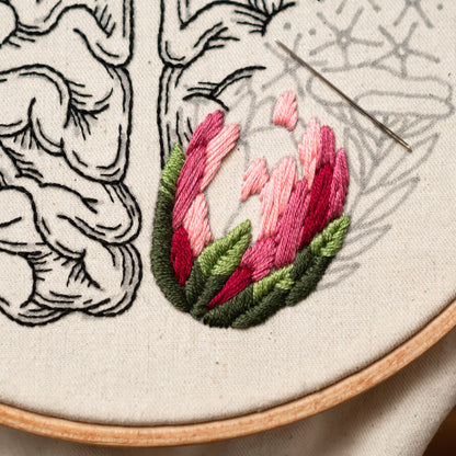 May include: Close-up of a hand-stitched embroidery design on a white fabric background. The design features a pink and green flower with a detailed outline of a brain in the background.
