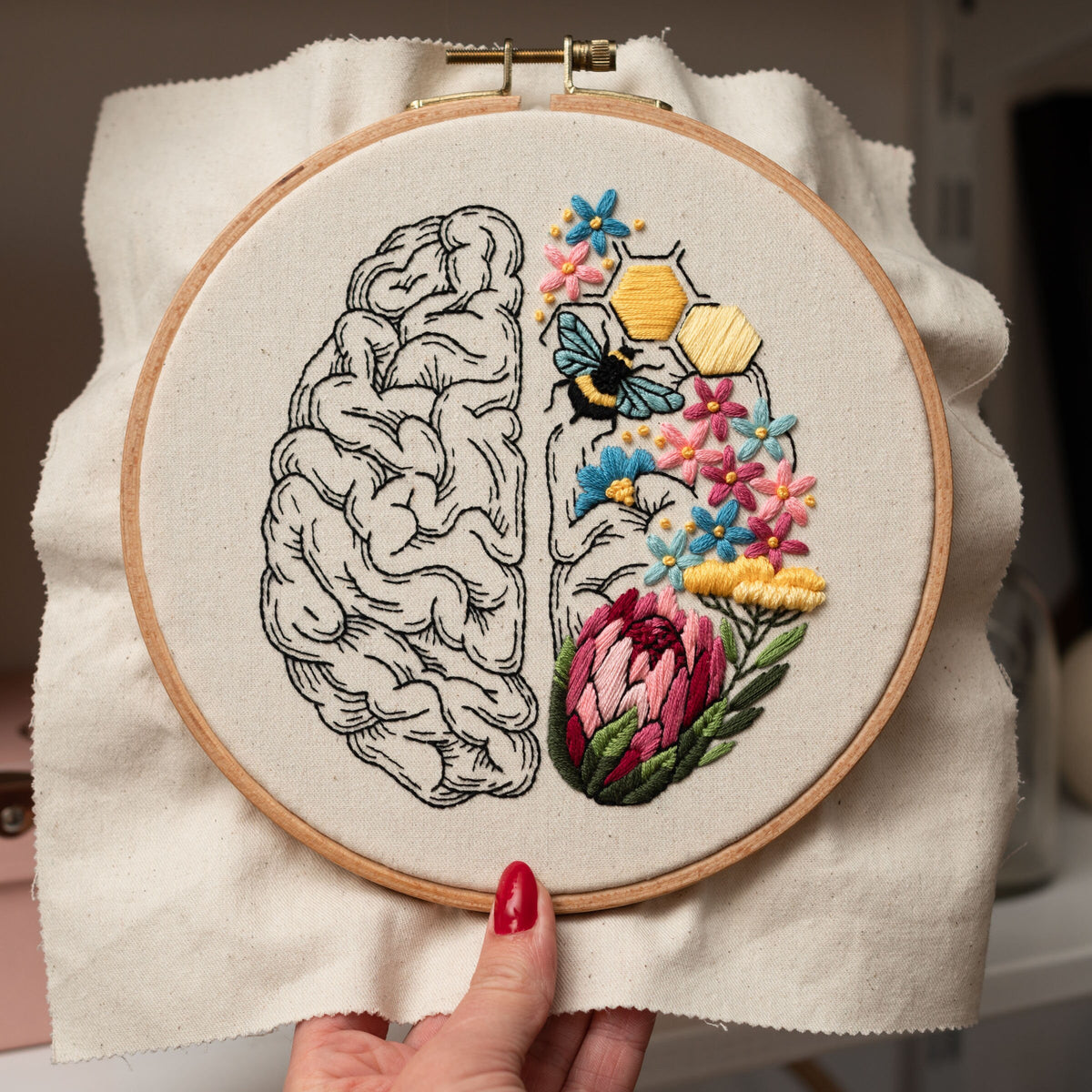 May include: Embroidered hoop art featuring a detailed outline of a human brain with a colorful floral design on one side and a bee and honeycomb on the other side.
