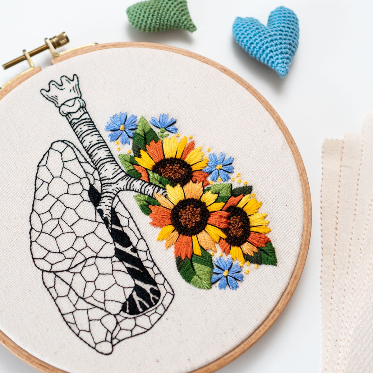 May include: Embroidered hoop art featuring a detailed anatomical illustration of a lung with a cluster of colorful sunflowers, blue and yellow wildflowers, and green leaves.
