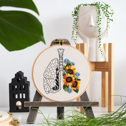 May include: Embroidered hoop art featuring a black line drawing of a human lung with a bouquet of sunflowers and blue flowers in the center of the lung.