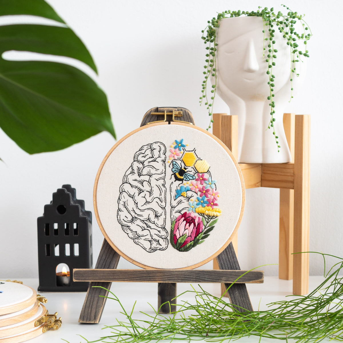 May include: Embroidered hoop art featuring a stylized brain with one half filled with colorful flowers and a bee, and the other half left blank. The hoop is on a wooden easel.