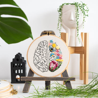 May include: Embroidered hoop art featuring a stylized brain with one half filled with colorful flowers and a bee, and the other half left blank. The hoop is on a wooden easel.
