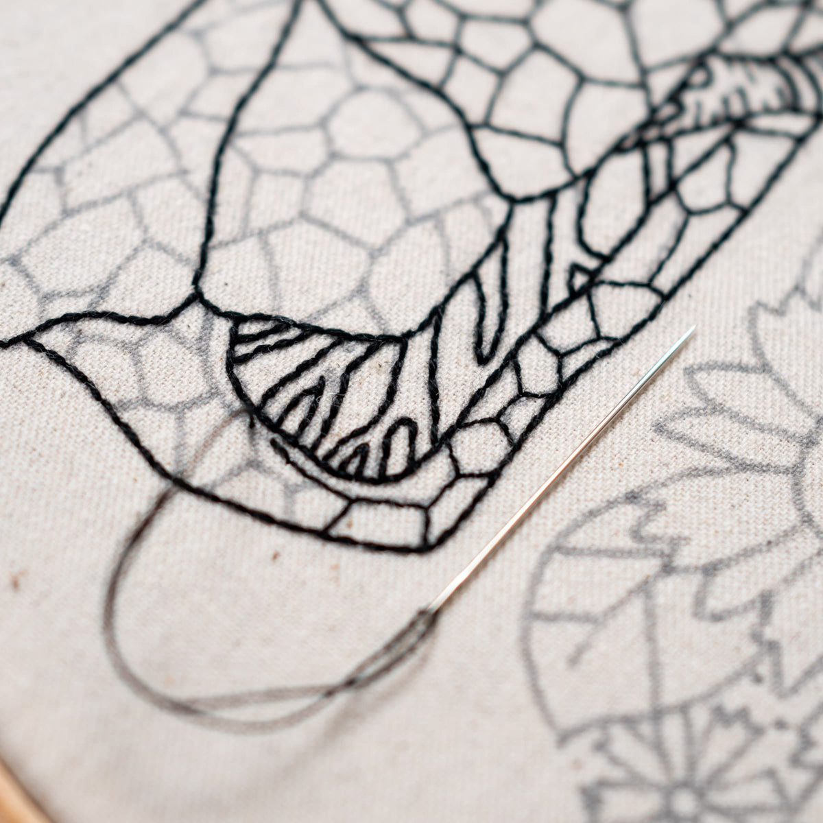 May include: Close-up of a needle and thread being used to embroider a black and white geometric design on a white fabric. The design features a series of interconnected shapes and lines.