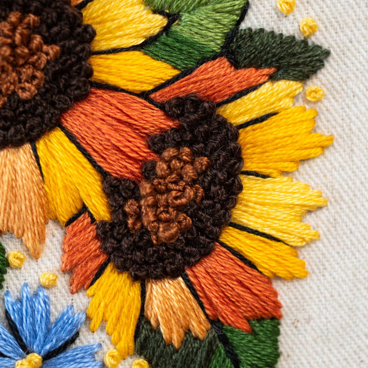 May include: Close-up of a hand-stitched embroidery design featuring multiple sunflowers with yellow, orange, and brown thread. The sunflowers are stitched on a white fabric background.