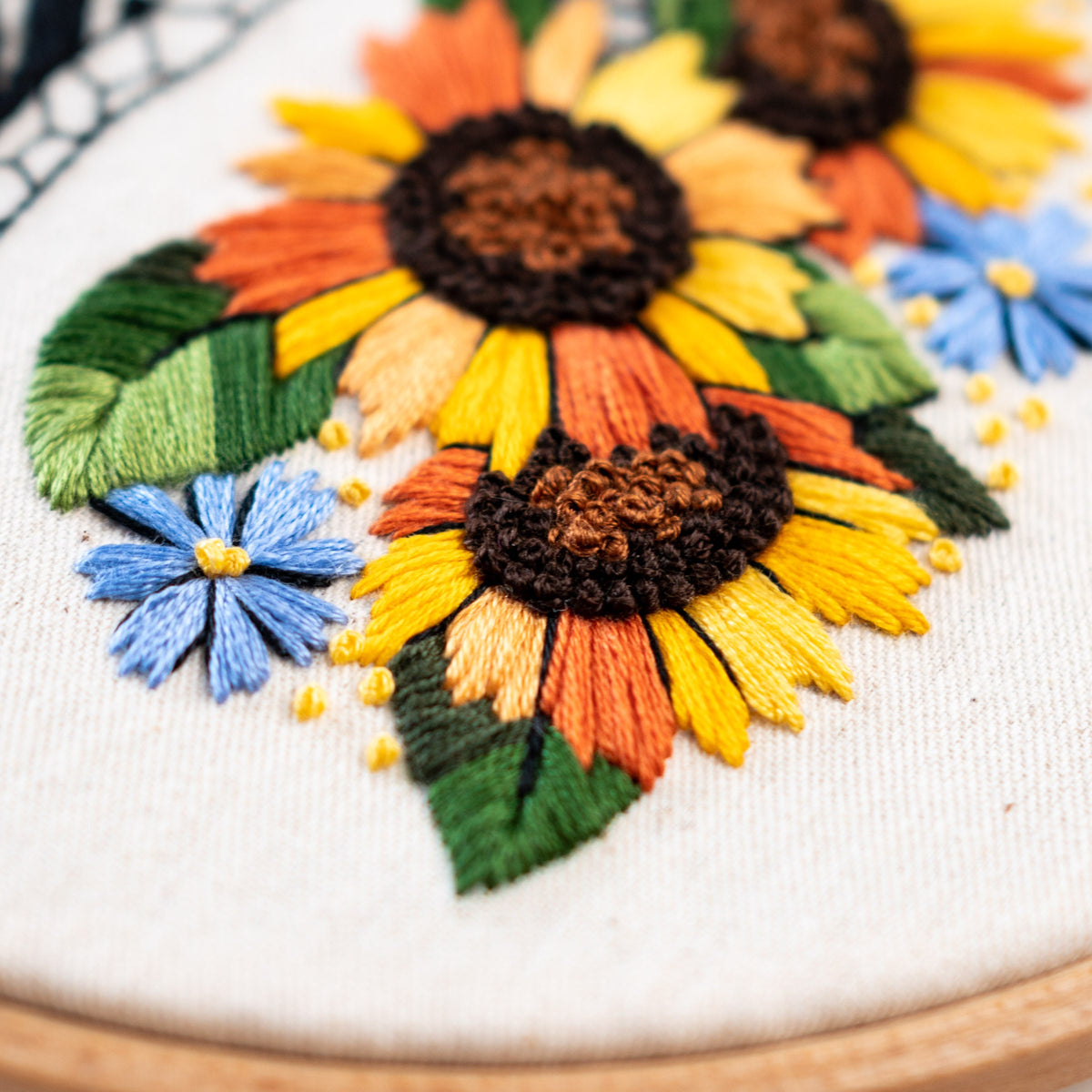 May include: Close-up of an embroidered hoop art piece featuring a cluster of yellow, orange, and brown sunflowers with blue and white wildflowers. The embroidery is done on a white fabric background.