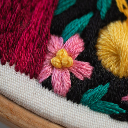 May include: Close-up of a pink and yellow embroidered flower on a black fabric background. The flower is surrounded by green and red embroidered leaves.