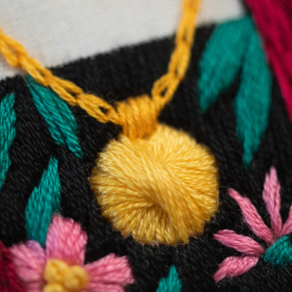 May include: Close-up of a yellow embroidered flower with green leaves and pink flowers on a black fabric background. The yellow flower is a knotted design and is part of a necklace.