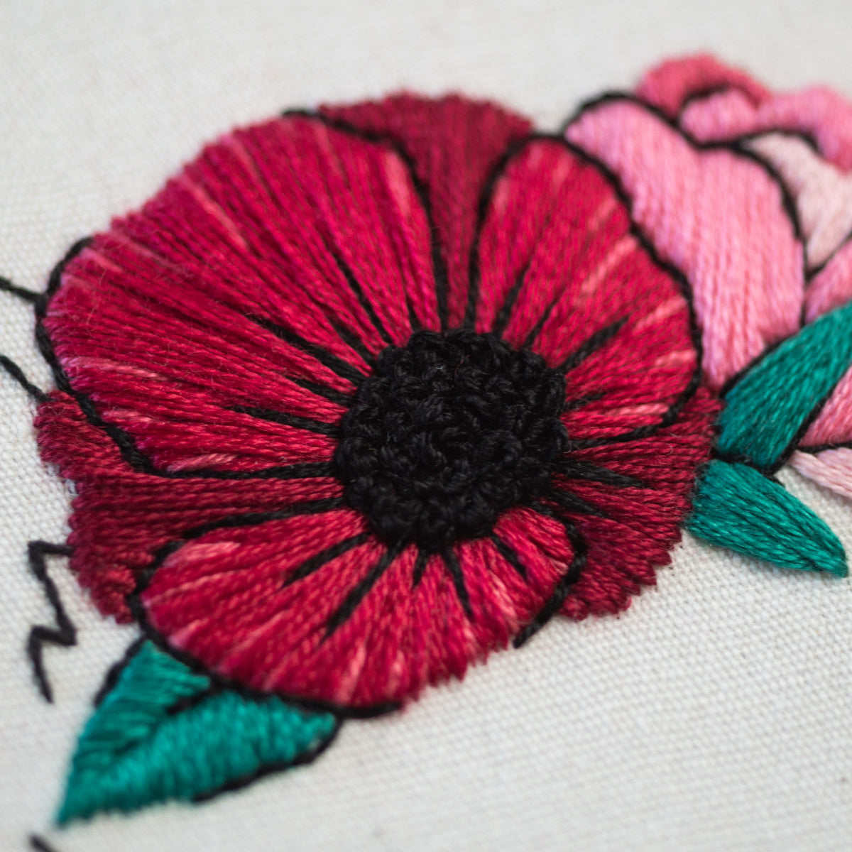 May include: Close-up of a red embroidered poppy flower with a black center and green leaves. The flower is stitched on a white fabric.