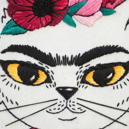 May include: Embroidered white canvas with a black and white cat face. The cat has yellow eyes, black eyebrows, and a black nose. The cat is wearing a red, pink, and green flower crown.