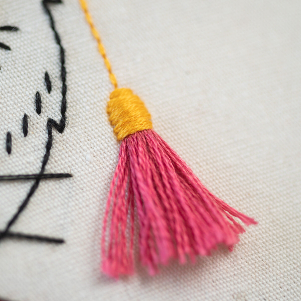 May include: A pink and yellow tassel with a yellow thread attached to the top. The tassel is made of soft, fluffy fibers and is attached to a white fabric background.