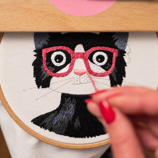 May include: Embroidered black and white cat with pink glasses and a pink nose. The cat is wearing a black collar and is being stitched with pink thread.