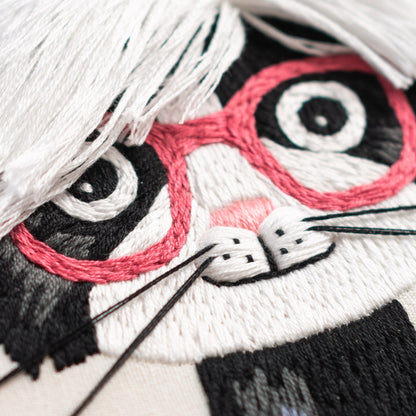 May include: Close-up of an embroidered white cat with black and pink details. The cat is wearing pink glasses and has a white nose and whiskers.