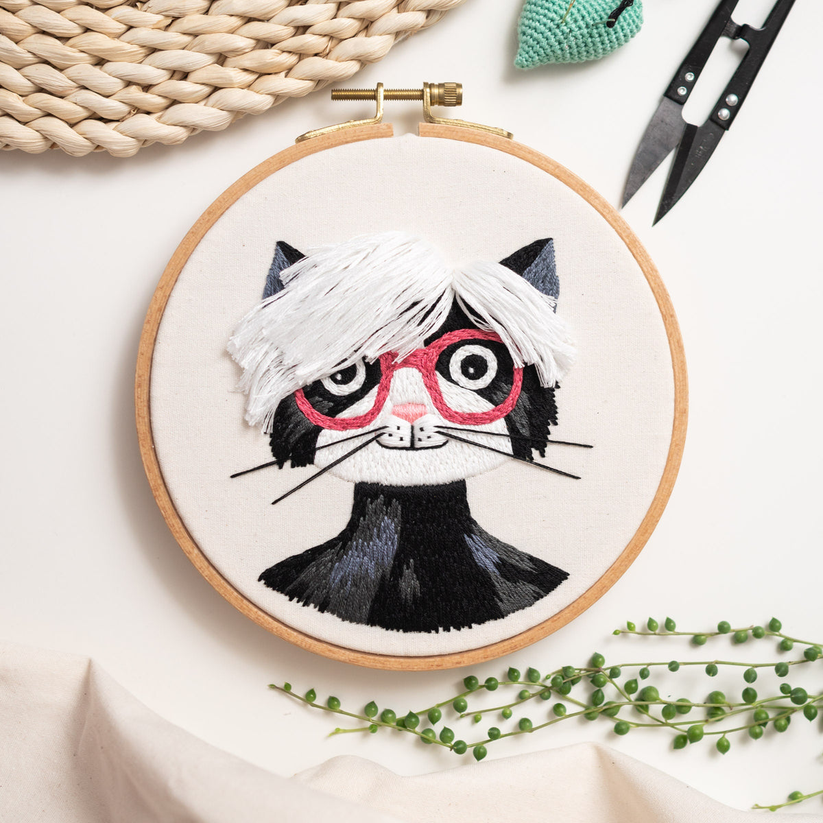 May include: Embroidered hoop art featuring a black and white cat with pink glasses and a white fluffy hairstyle. The cat is embroidered on a white fabric background.