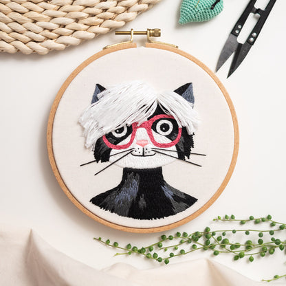 May include: Embroidered hoop art featuring a black and white cat with pink glasses and a white fluffy hairstyle. The cat is embroidered on a white fabric background.