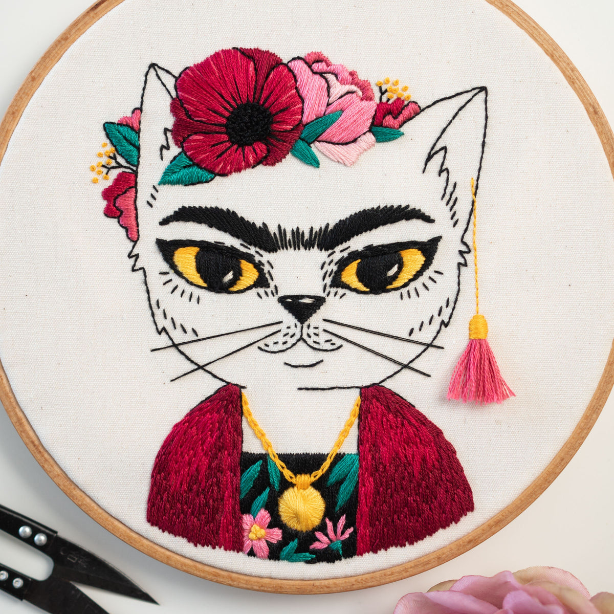 May include: Embroidered white cat with a red and pink flower crown, a red jacket, and a gold necklace. The cat has black eyes and a black nose. The background is white.