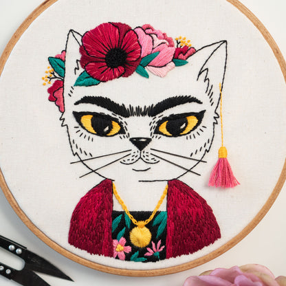 May include: Embroidered white cat with a red and pink flower crown, a red jacket, and a gold necklace. The cat has black eyes and a black nose. The background is white.