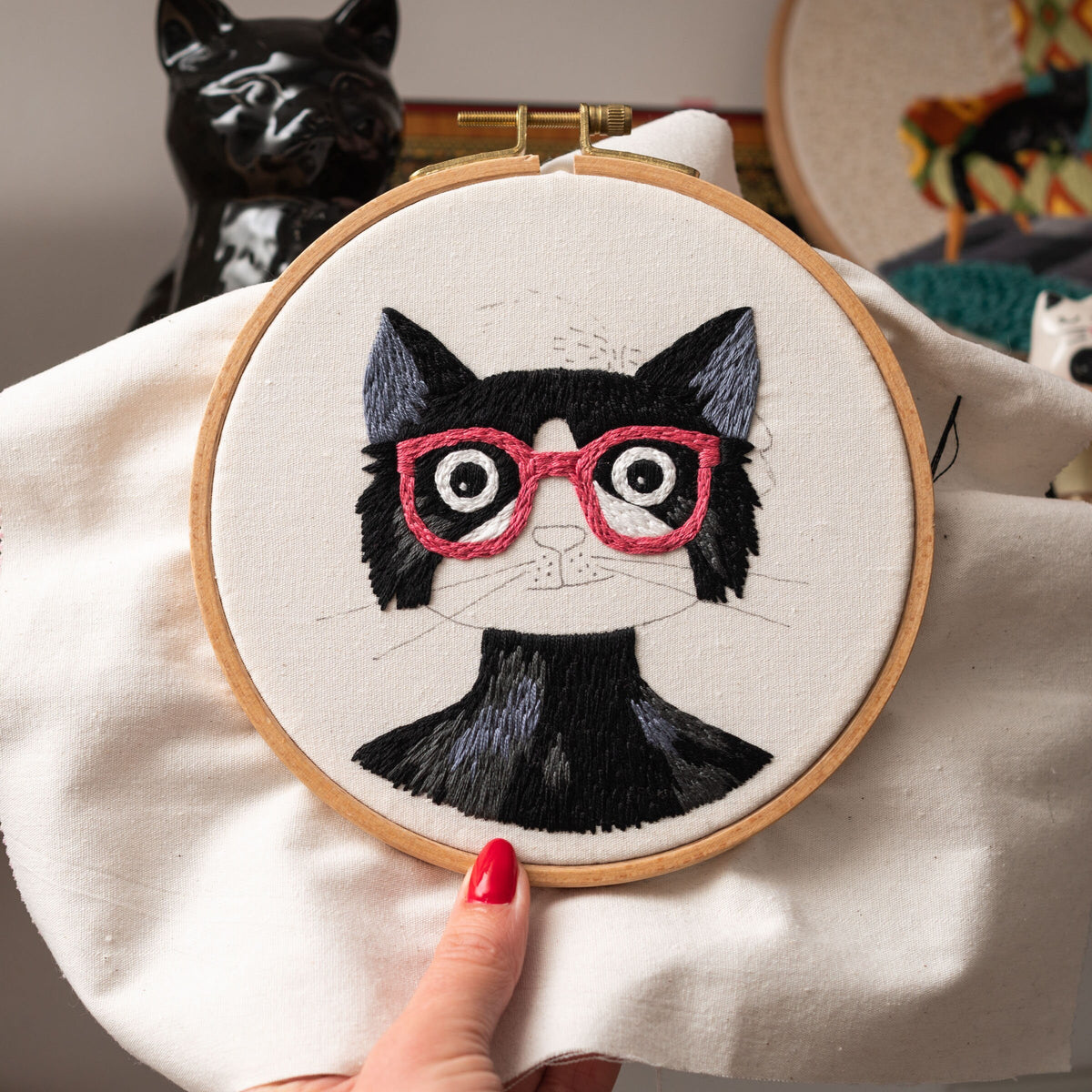 May include: Embroidered black cat with pink glasses on a white fabric hoop. The cat has a black nose and white whiskers.