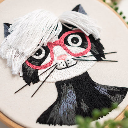 May include: Embroidered black and white cat with pink glasses and a white fringe hairstyle. The cat is stitched on a white canvas.