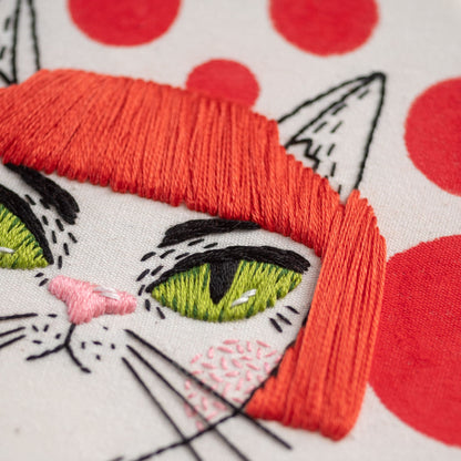 May include: Close-up of an embroidered cat with green eyes, a pink nose, and a red fringe. The cat is on a white background with red polka dots.