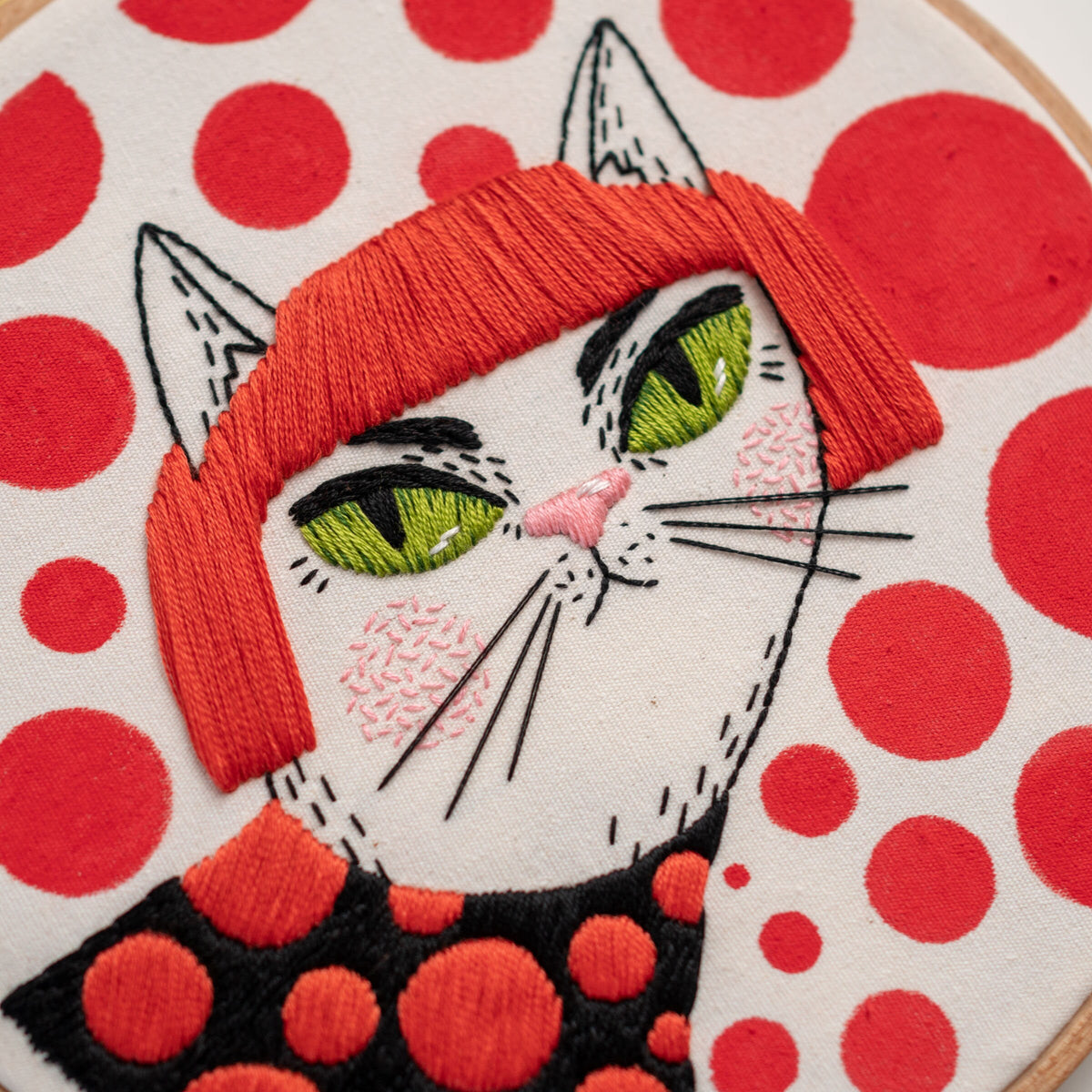 May include: Embroidered white cat with red hair and green eyes, wearing a black and red polka dot shirt. The background is white with red polka dots.